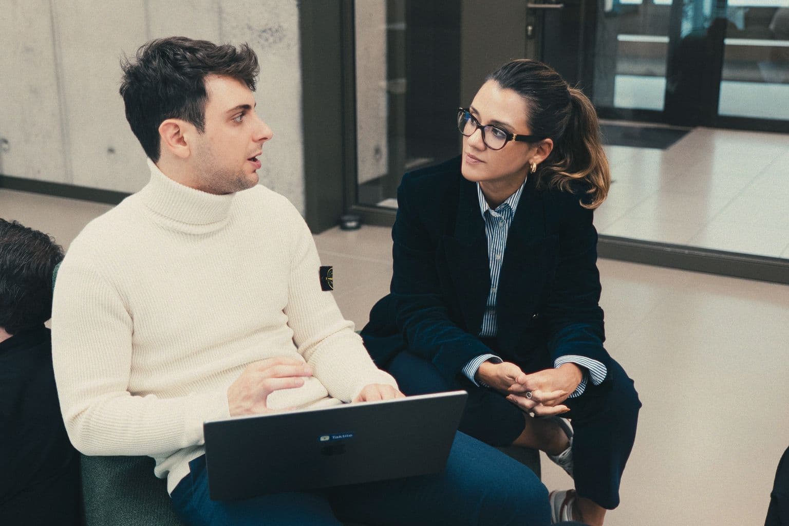 Two colleagues in conversation in an office lounge, one holding a laptop